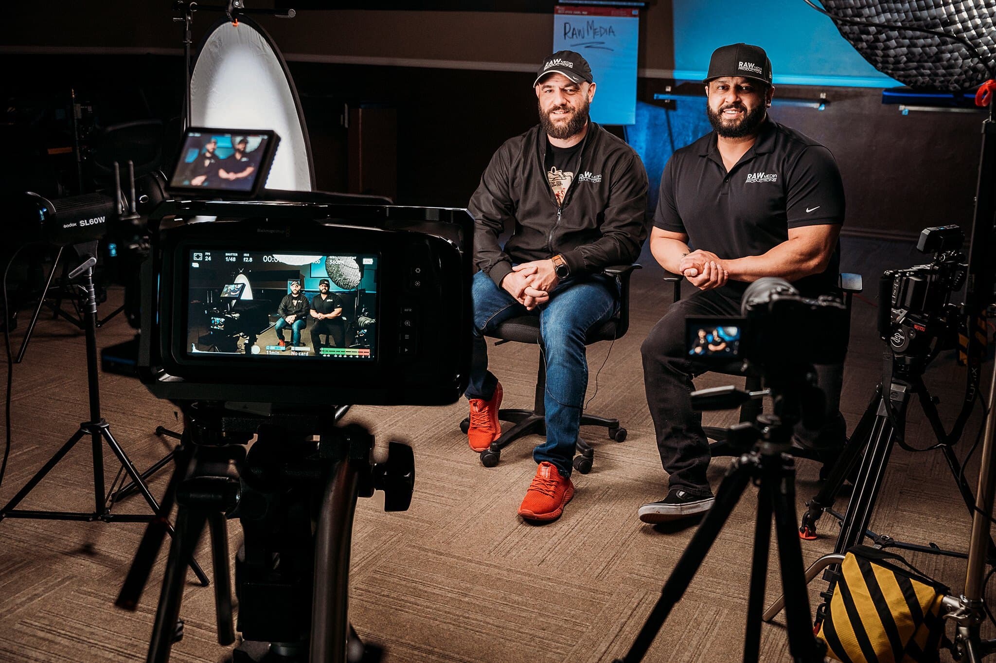 Raw Media Pros founders seated on set beside cameras and monitors